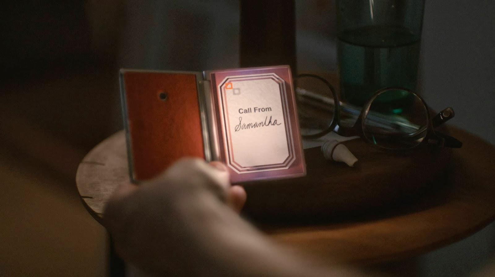 Theodore's handheld device from the film Her resting on a nightstand, its screen reading Call From Samantha, beside a pair of glasses and an earpiece